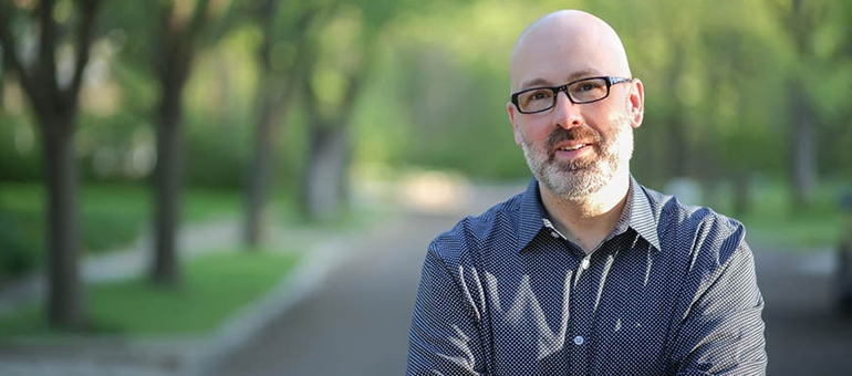 Sean Goggins stands in a road in front of lawns and trees while wearing a grey shirt.