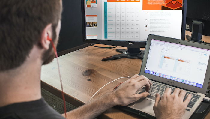 Man wearing headphones works on laptop with desktop monitor behind him