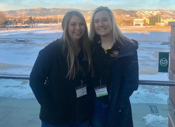 Two women stand in front of a snowy field with mountains in the background.